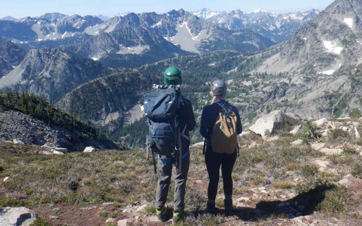 two people wearing helmets and backpack look out over a vast mountain landscape